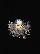 A handcrafted silver brooch features an intricate white rose on a blue backdrop, accented with delicate silver leaves and small round details over a black background, echoing the elegance of CAMELLIA's Cotton Rose Hibiscus Birth Flower Ring.